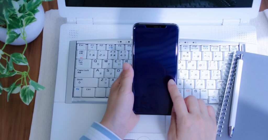 ノートパソコンの上でスマートフォンを操作する手元